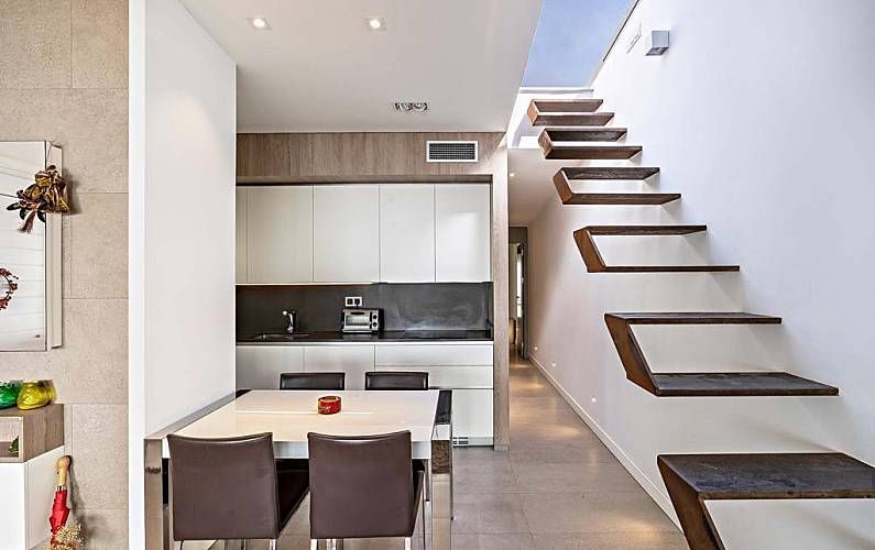 Cocina moderna con escalera abierta, armarios blancos, mesa de comedor y encimeras de color gris oscuro.