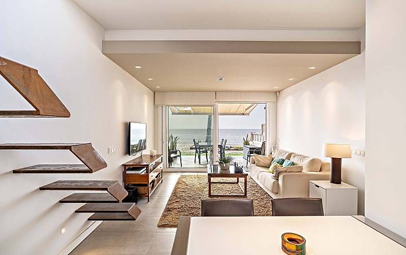 Salón moderno con vistas al océano, paredes blancas, escalera flotante de madera y grandes puertas correderas de cristal.