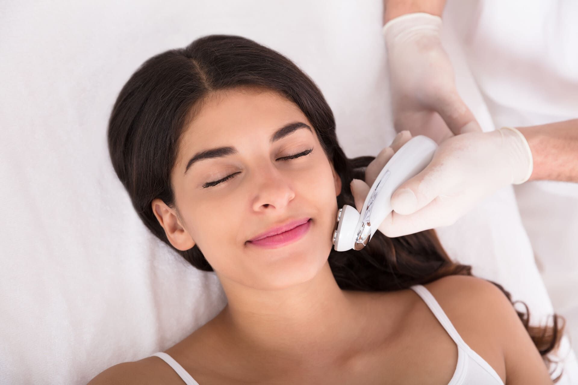 Una persona, con los ojos cerrados, recibe un tratamiento facial profesional mediante un dispositivo portátil en un entorno clínico.