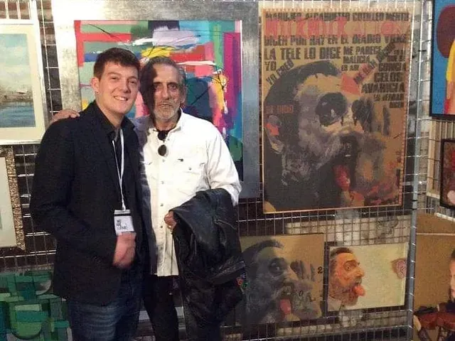Dos hombres posan para una foto frente a una exposición de pinturas.