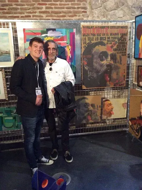 Dos hombres posan para una fotografía delante de una pared de pinturas.