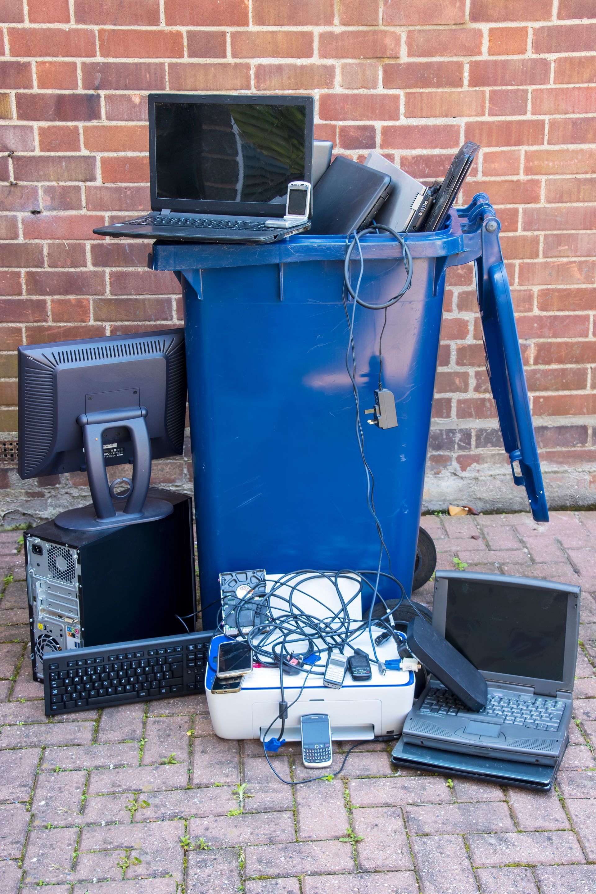 Une poubelle bleue débordant de déchets électroniques est adossée à un mur de briques.