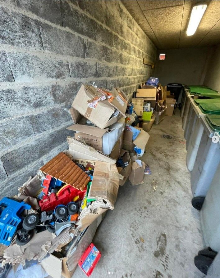 Des cartons d'ordures et de jouets s'entassent contre un mur de briques, à côté de poubelles vertes et grises.