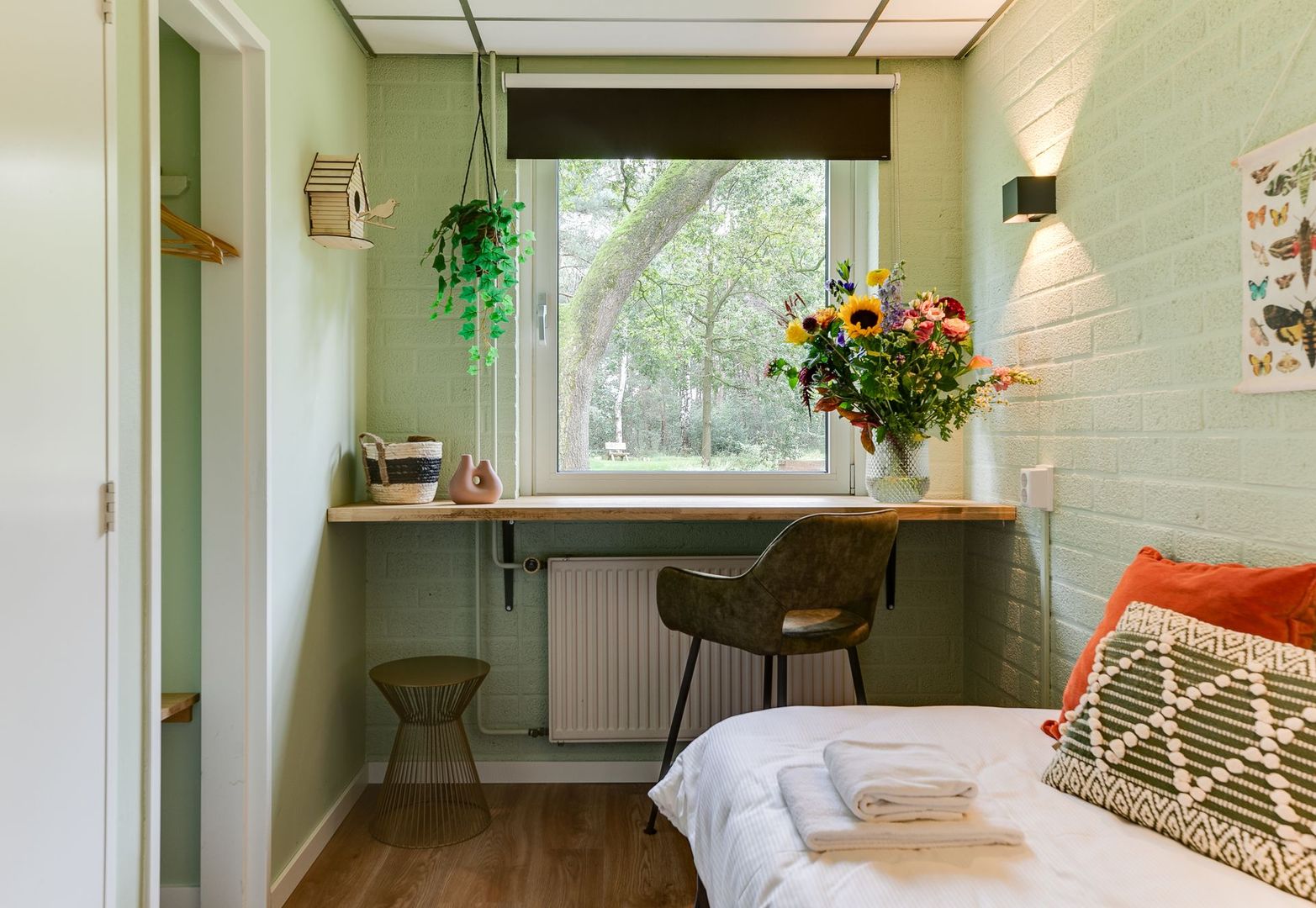 Slaapkamer met bureau bij het raam, mintgroene muren, houten vloer, zonnebloemen, wit bed en stoel.
