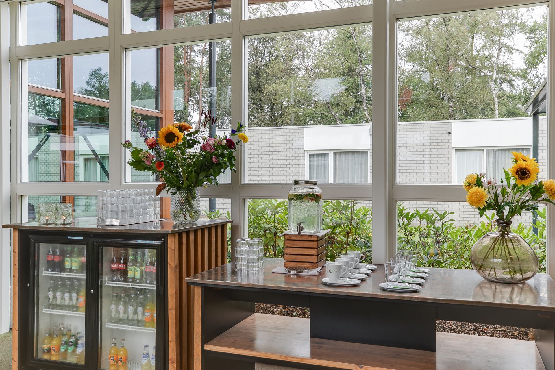 Bargedeelte met drankjes en bloemen bij grote ramen, met uitzicht op een groene buitenruimte.