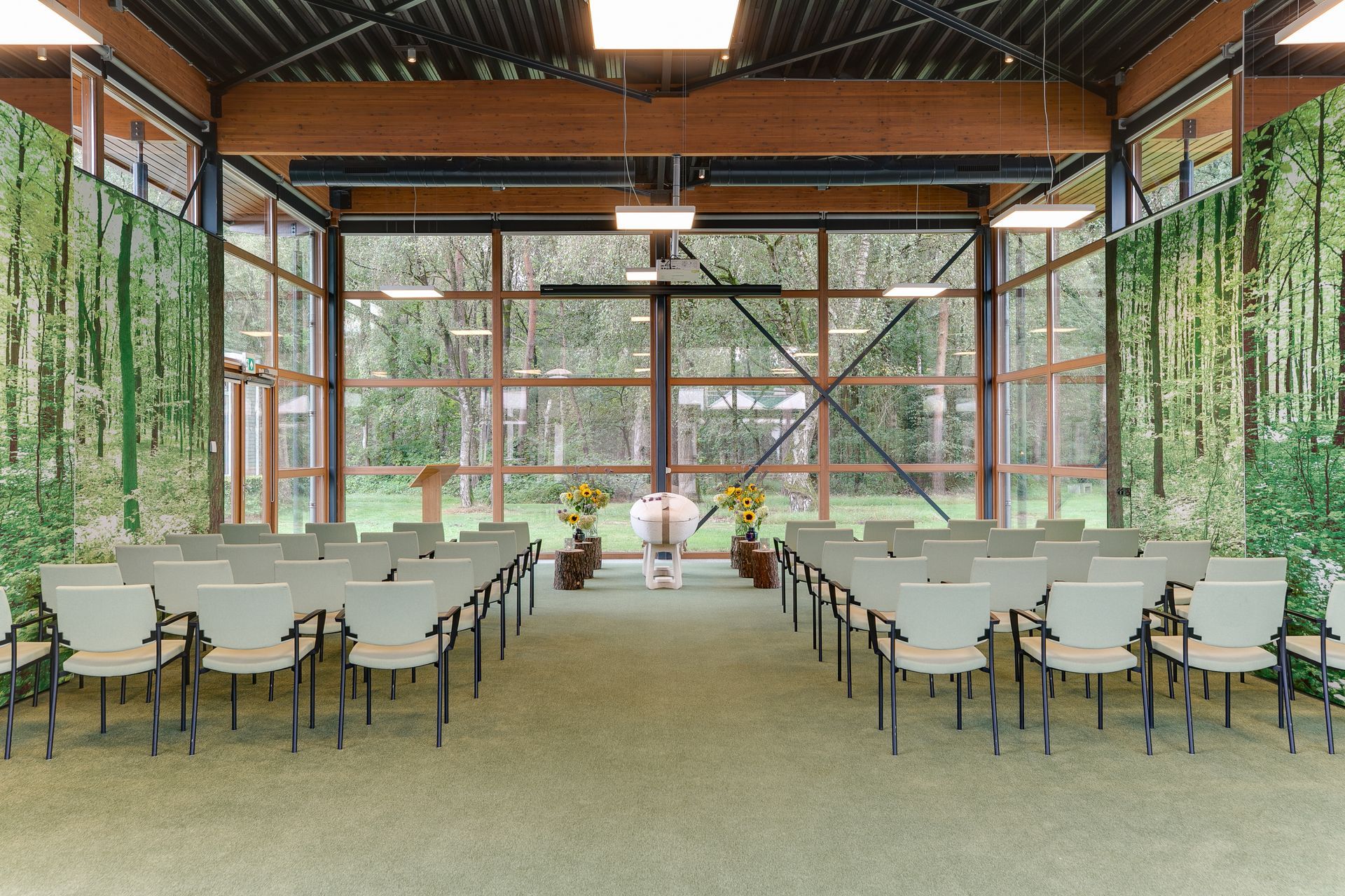 Een evenementenhal met rijen stoelen tegenover een bloemstuk voor een raam met een muurschildering van een bos.