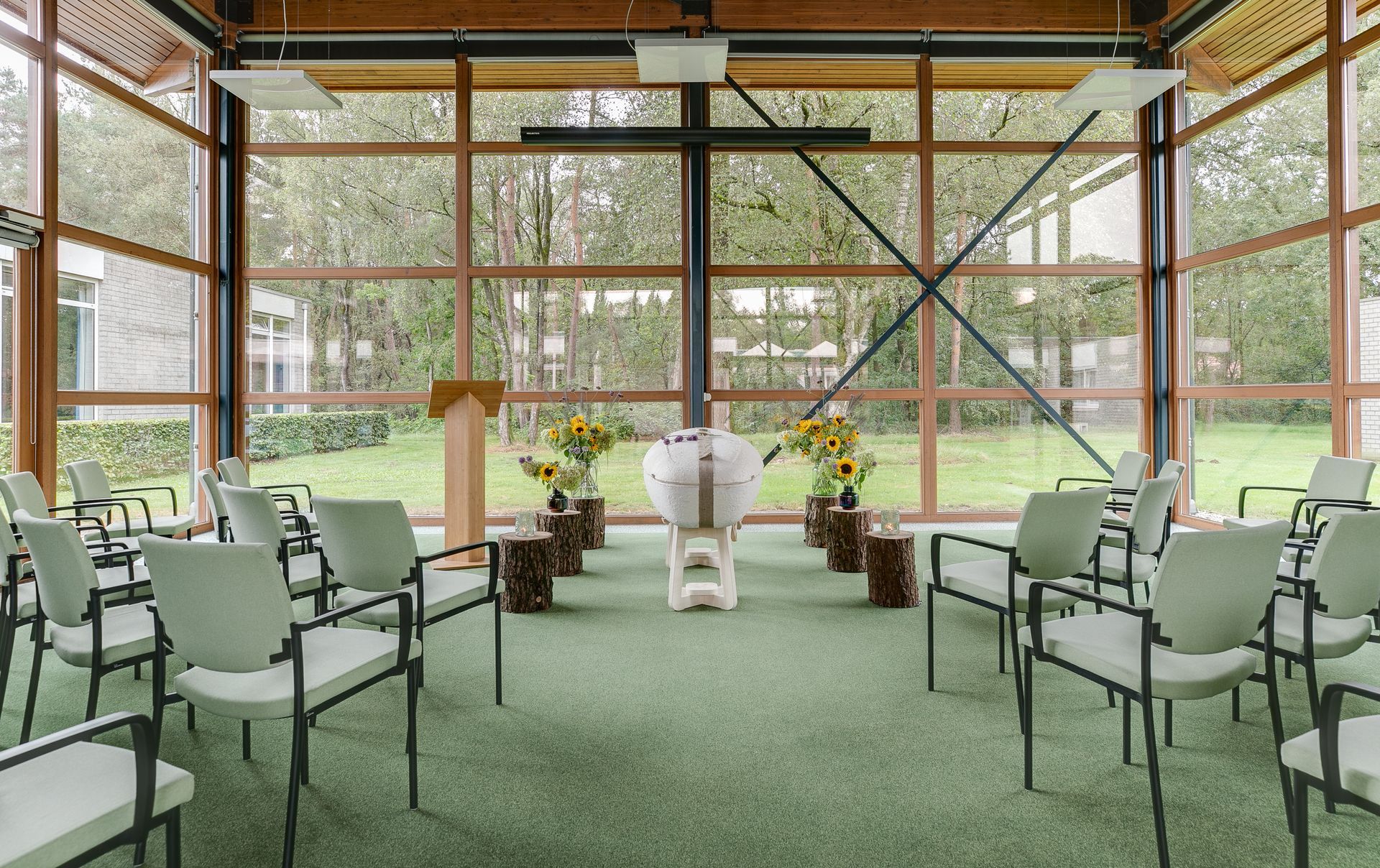 Interieur van een kapel met glazen wanden, stoelen, een podium, bloemstukken en een centraal altaar. Groen tapijt en uitzicht naar buiten.