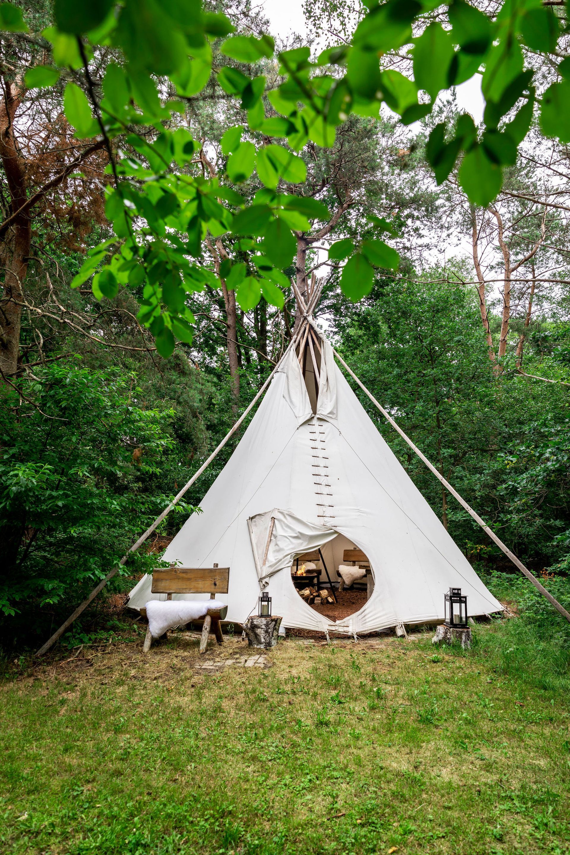 Witte tipi in een groene, bosrijke omgeving. Houten palen, ronde ingang en bord.