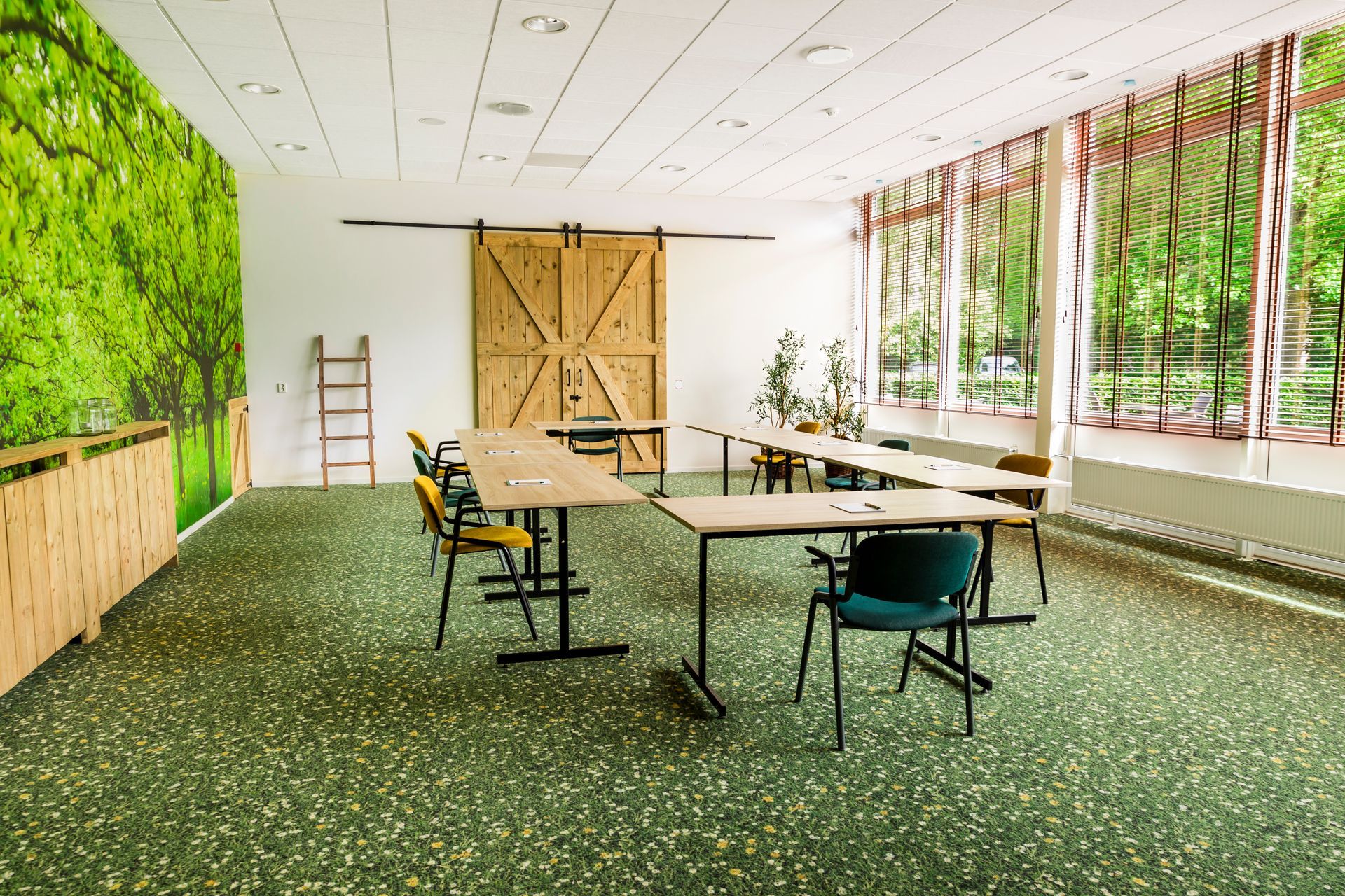 Klaslokaal met een groen gespikkelde vloer, tafels in een U-vorm, grote houten deuren en een muurschildering van bomen.