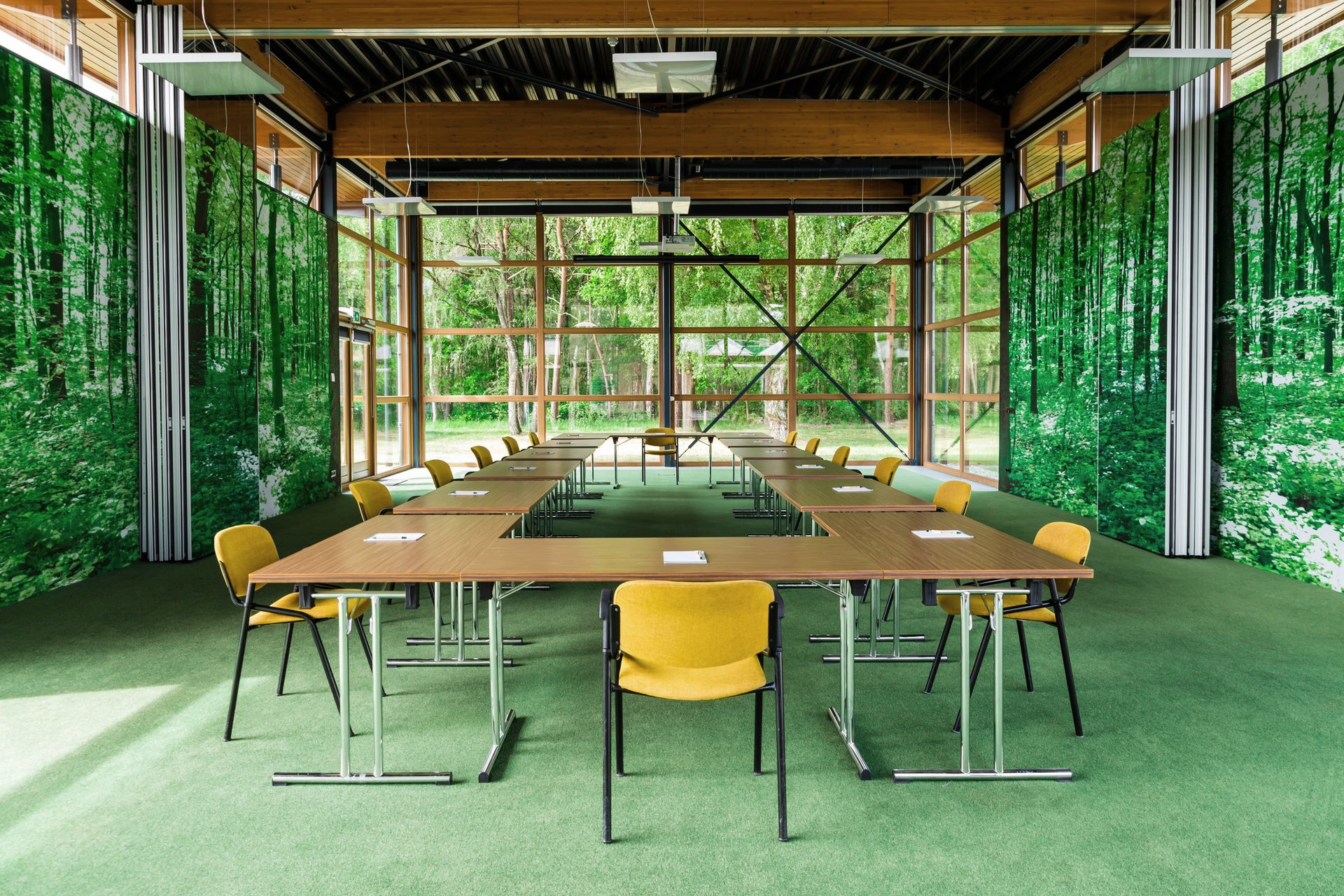 Vergaderruimte met U-vormige tafel en gele stoelen, groen tapijt en muurschilderingen van een bos.