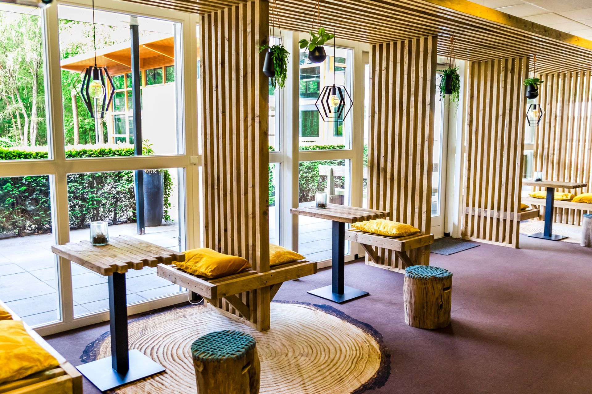 Een lichte, luchtige kamer met houten balken en ramen, kleine tafels en banken met gele kussens.