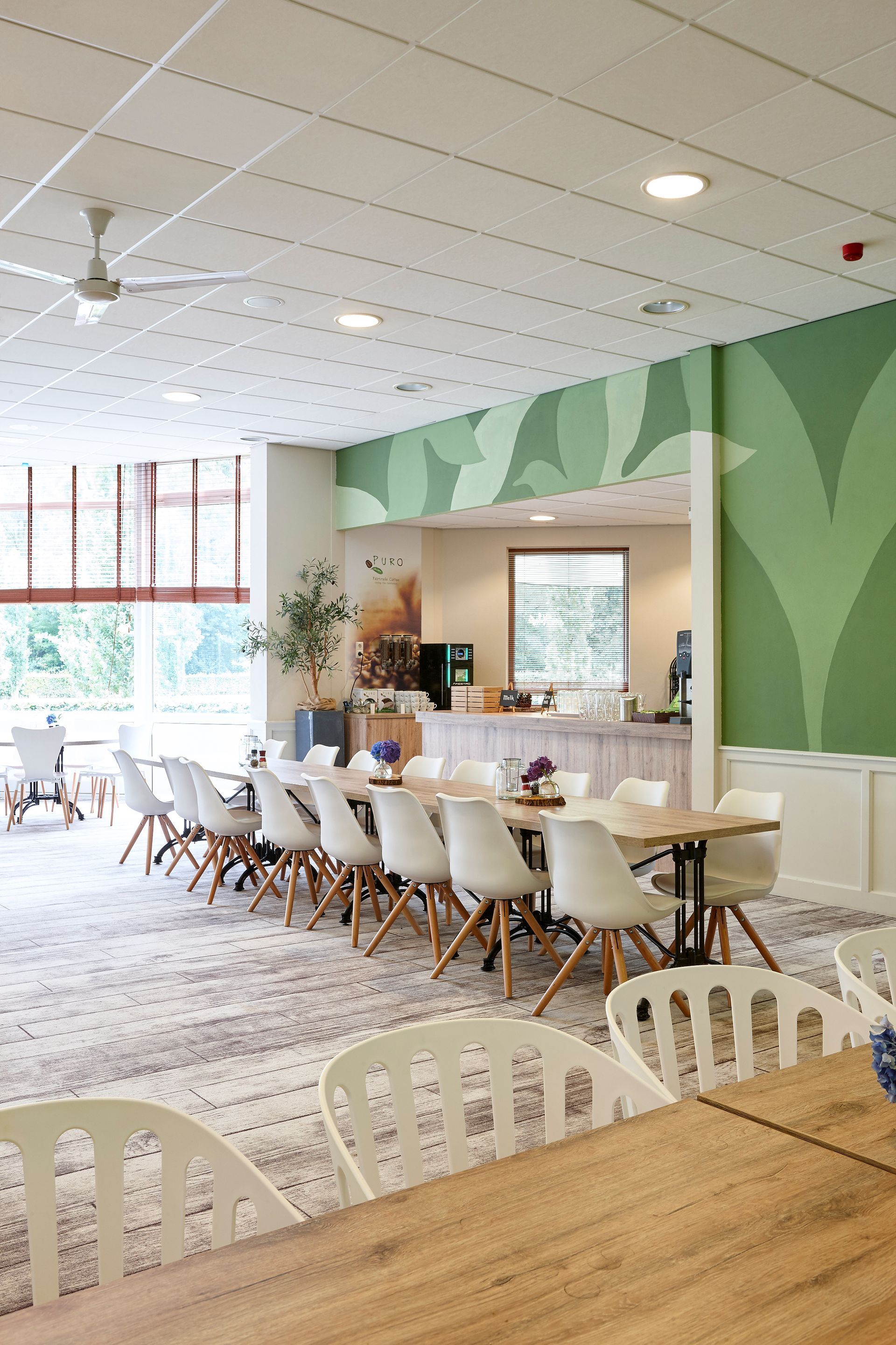 Eetkamer met lange tafel, witte stoelen, groene muurschildering en grote ramen.