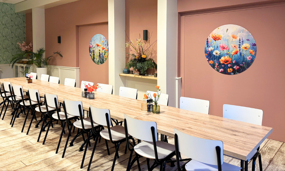 Lange houten tafel met witte stoelen in een kamer met roze muren, bloemstukken en decoratie.