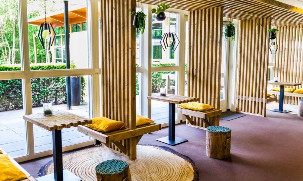 Een licht café met houten accenten en gele kussens, met tafels en een groot raam met uitzicht op het groen.