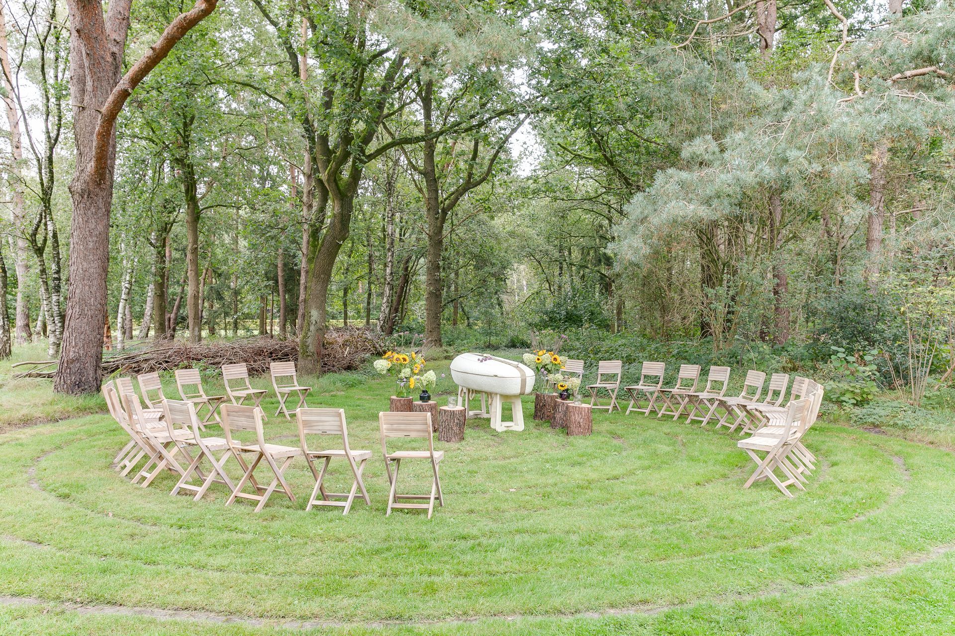 Een kring van houten stoelen op een grasveld, bomen op de achtergrond, een kleine bruiloftssetting.