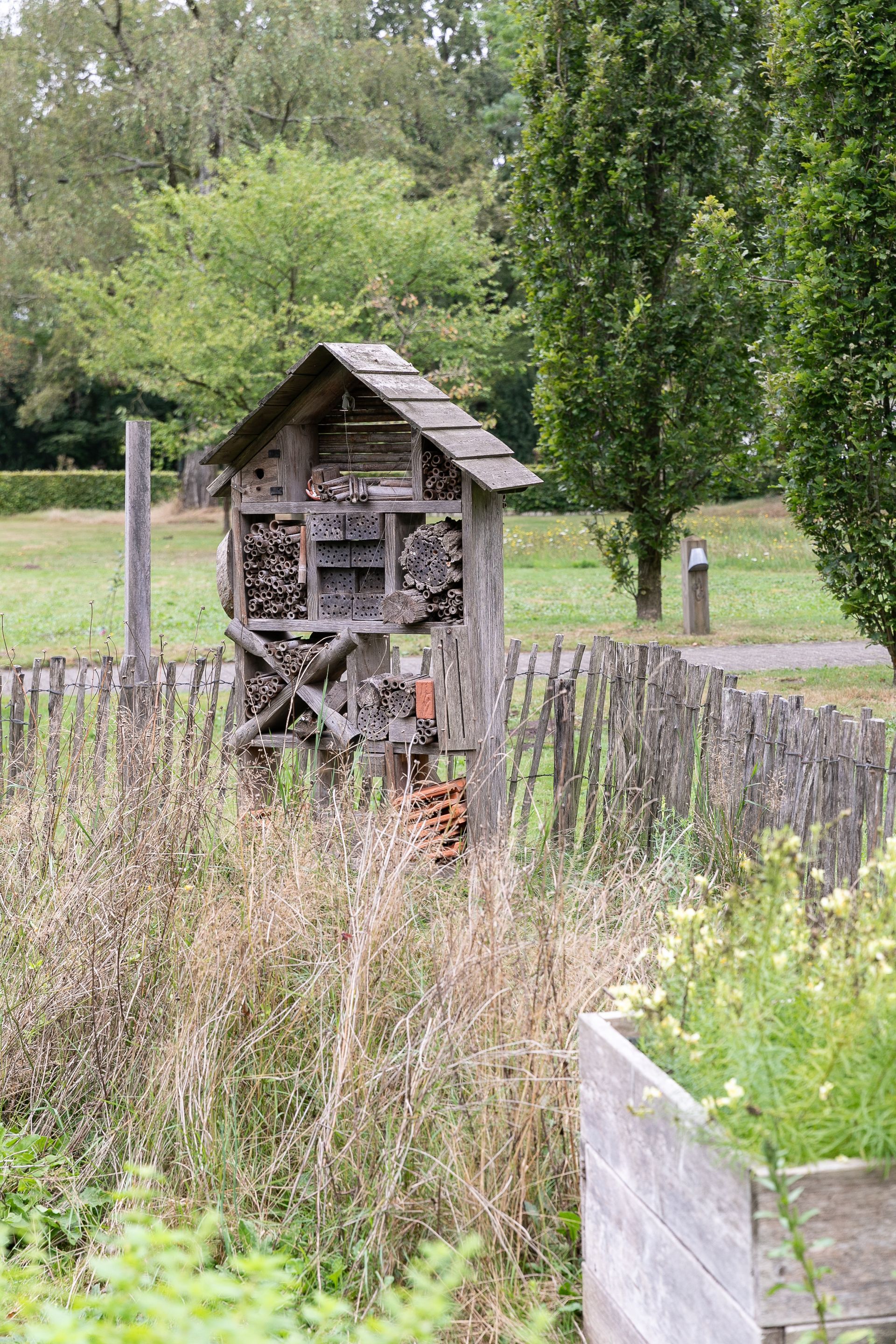 Houten insectenhotel in een tuin met een schutting en hoge bomen, omgeven door planten.