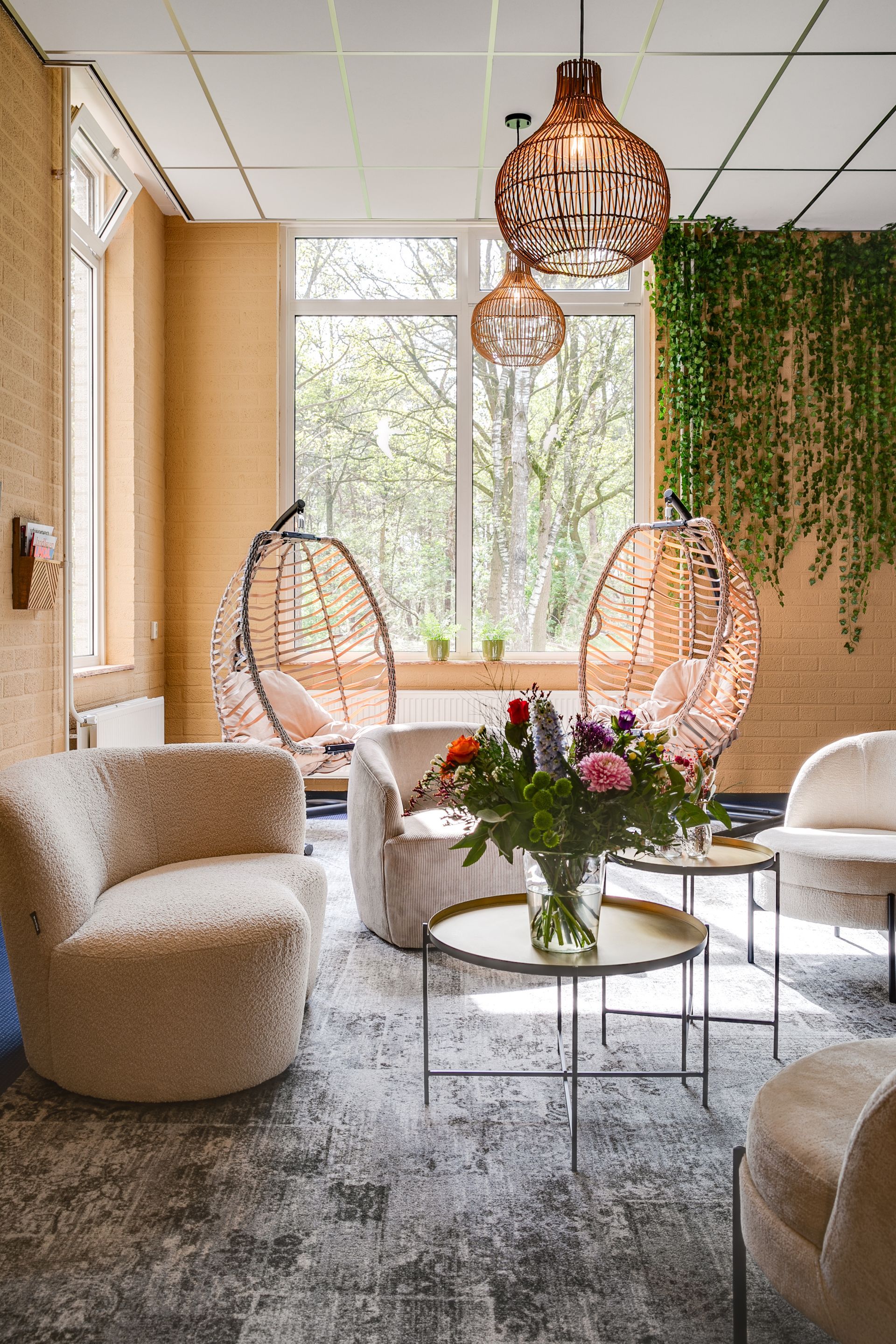 Gezellige zithoek met lichtgekleurde stoelen, hangende rieten zitjes en een groot raam met groen.