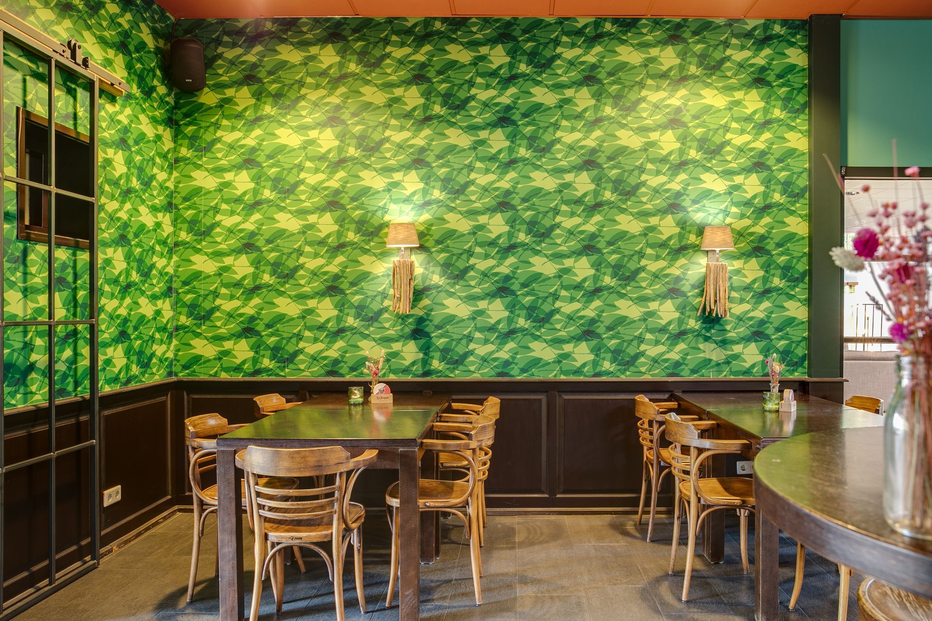Restaurantinterieur met behang met groene bladeren, houten tafels en stoelen en decoratieve wandlampen.