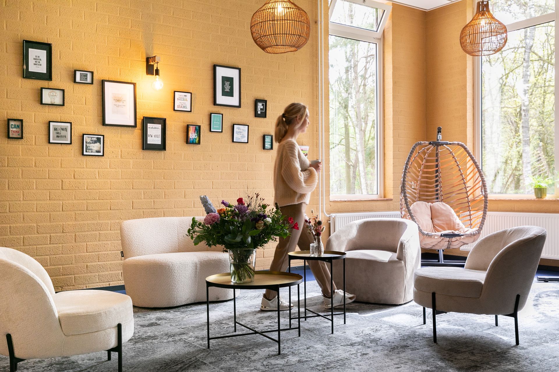 Vrouw in een knusse kamer met stoelen, schilderijen en een hangstoel, kijkt uit door een groot raam.