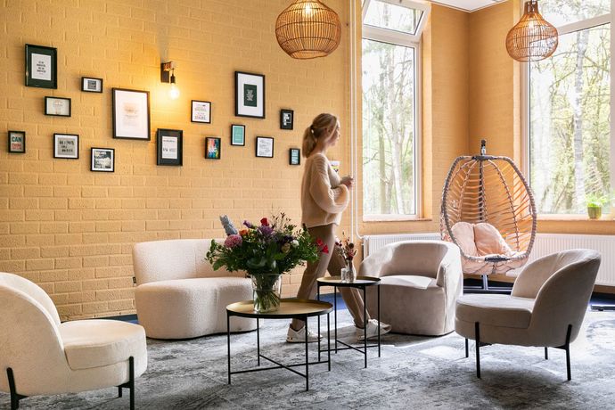 Vrouw in een knusse kamer met stoelen, schilderijen en een hangstoel, kijkt uit door een groot raam.