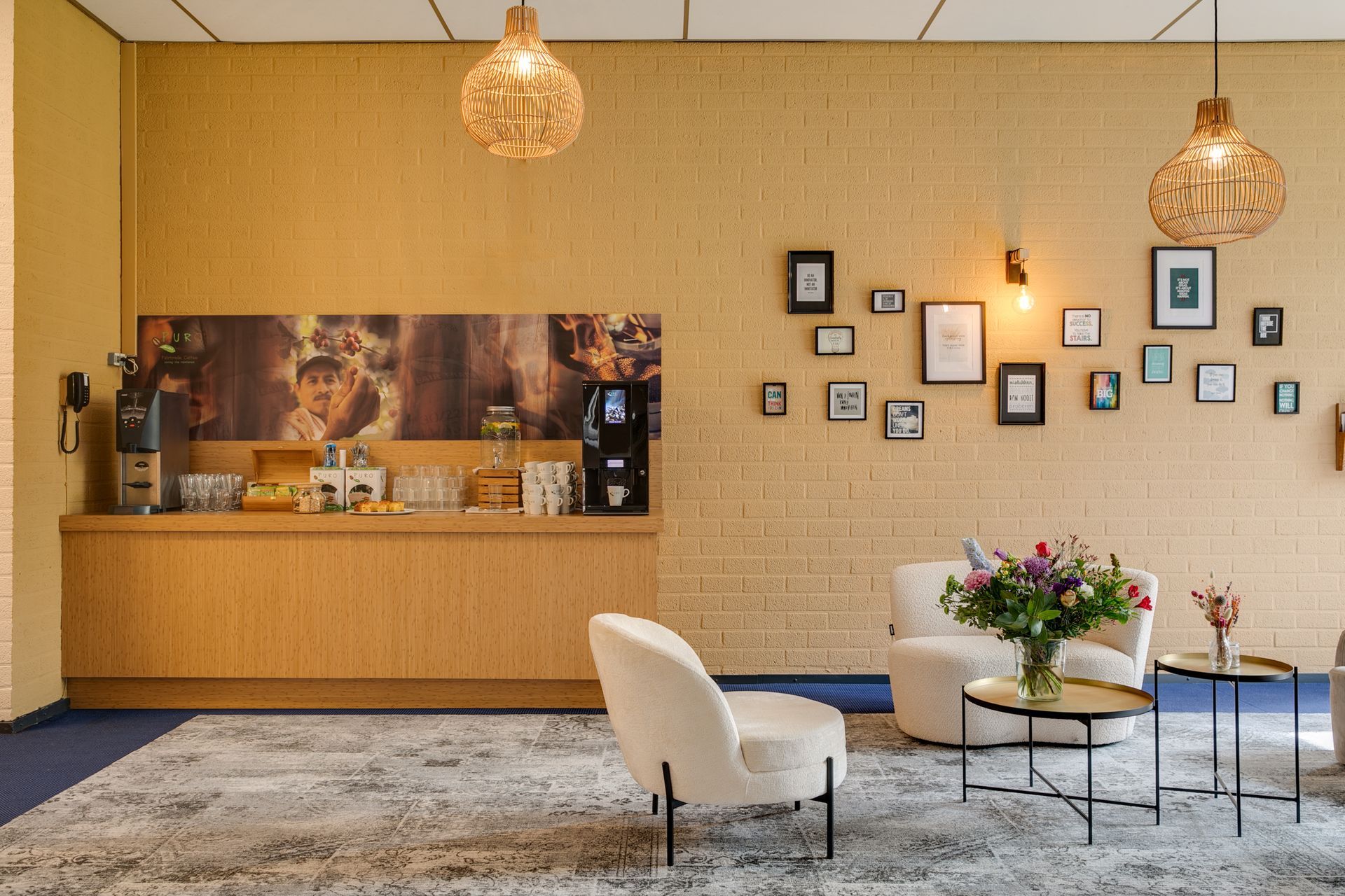 Gezellige kantoorlounge met koffiebar, fauteuils, bijzettafels en kunstwerken op een gele muur met structuurverf.
