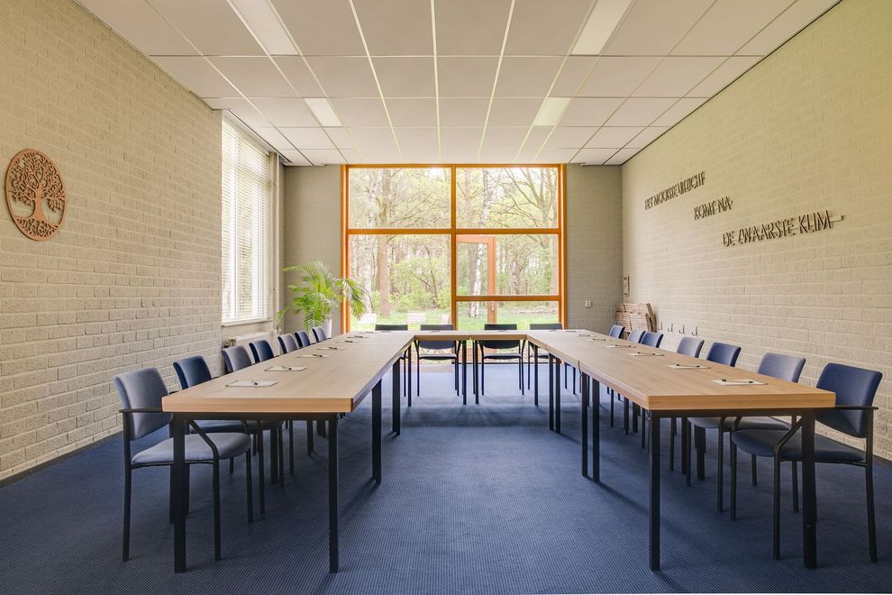 Vergaderruimte met een lange houten tafel, stoelen en grote ramen met uitzicht op bomen.