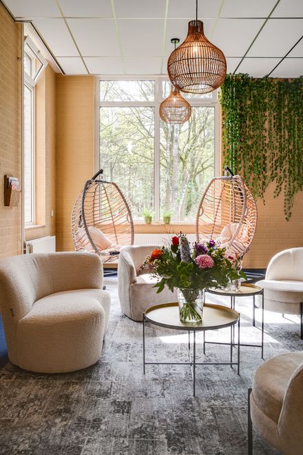 Gezellige zithoek met twee hangstoelen, banken en salontafels, vlakbij een raam met groen.