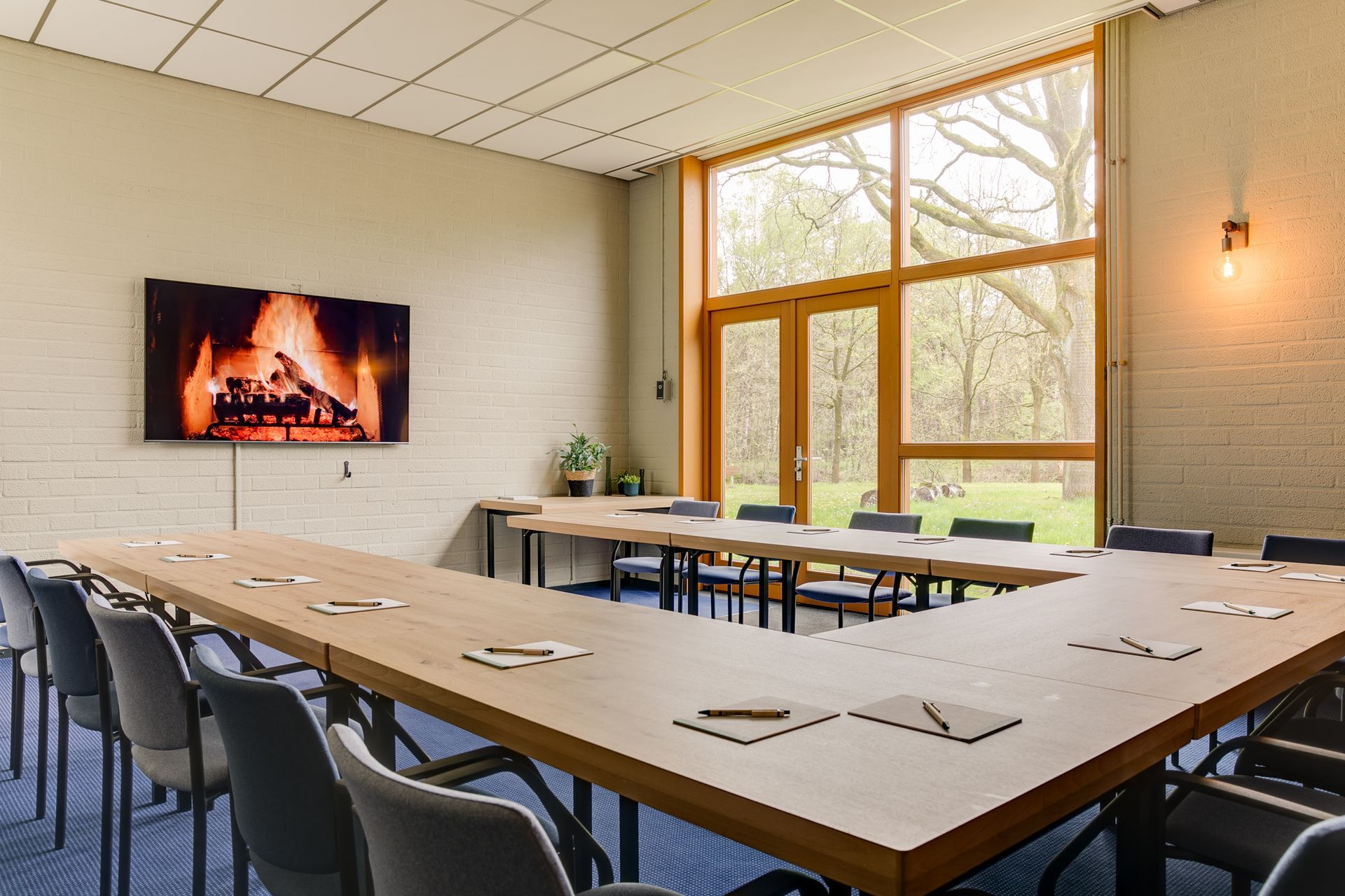 Vergaderruimte met U-vormige tafel, stoelen, tv met open haard en groot raam met uitzicht naar buiten.