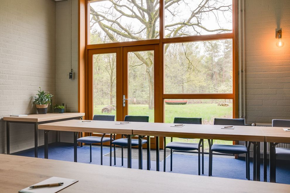 Vergaderruimte met een lange houten tafel, stoelen en grote ramen met uitzicht op bomen.