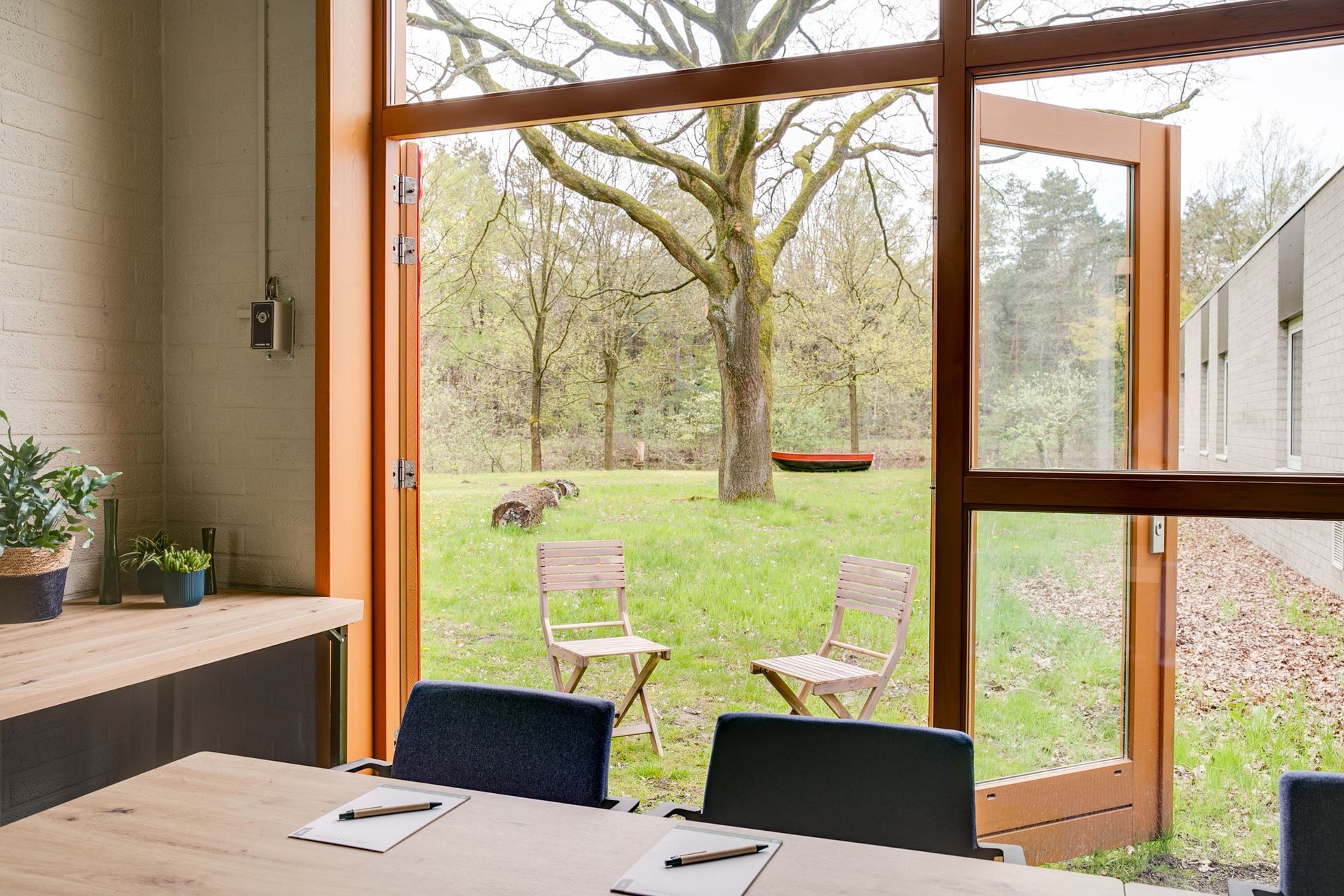 Open houten deuren geven toegang tot een grasrijke achtertuin met stoelen en een boom; binnen staan ​​een tafel en stoelen.