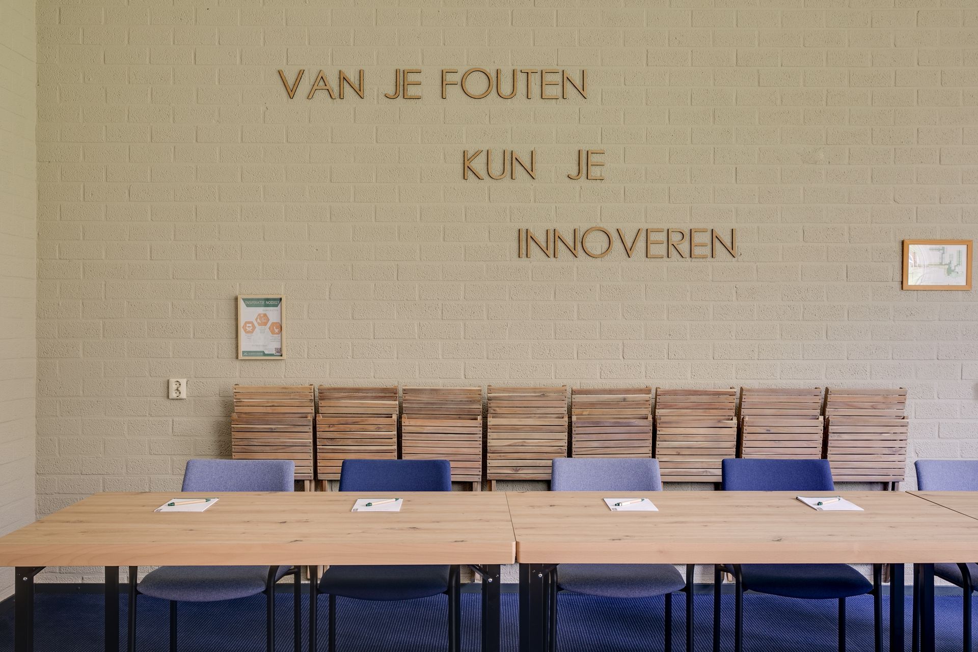 Vergaderruimte met een lange houten tafel, blauwe stoelen en een muur met de Nederlandse tekst: "Van je fouten kun je innoveren."
