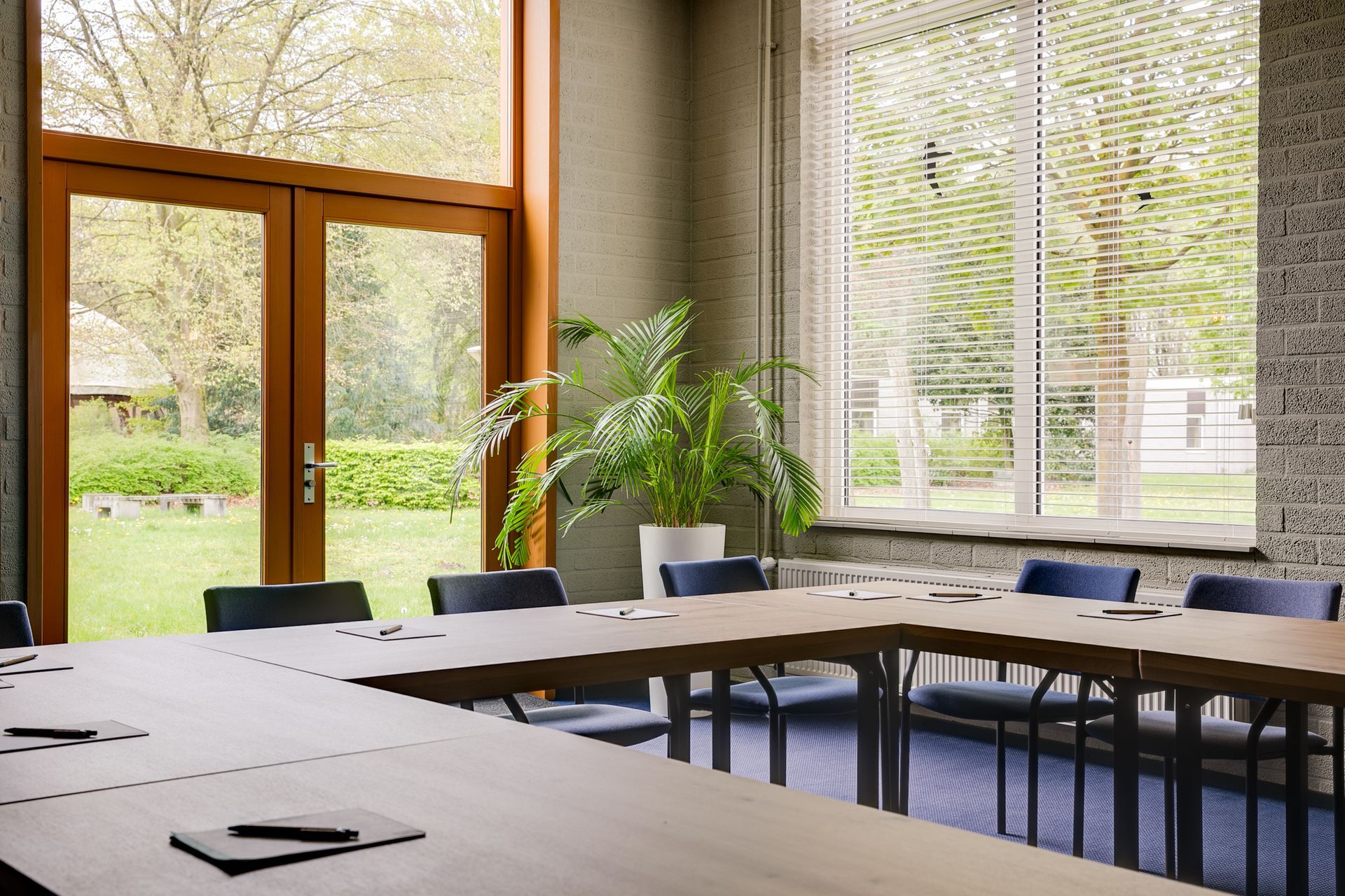 Vergaderruimte met U-vormige tafel, gele stoelen, groen tapijt, muurschildering van een bos, glazen ramen en houten balken.