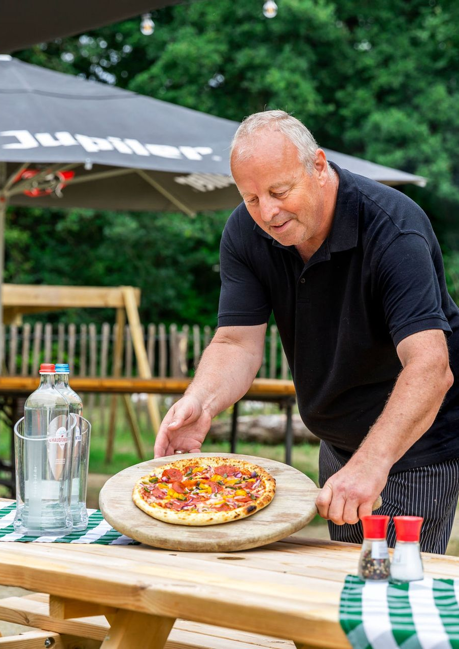 Een tuintafereel met een houten pergola, opgestapeld brandhout en een bijenkorf, te midden van weelderig groen.