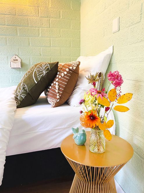 Bed met kussens, bloemen in een vaas en een gouden bijzettafel. Groengetinte muur, wit beddengoed en kleurrijke bloemen.