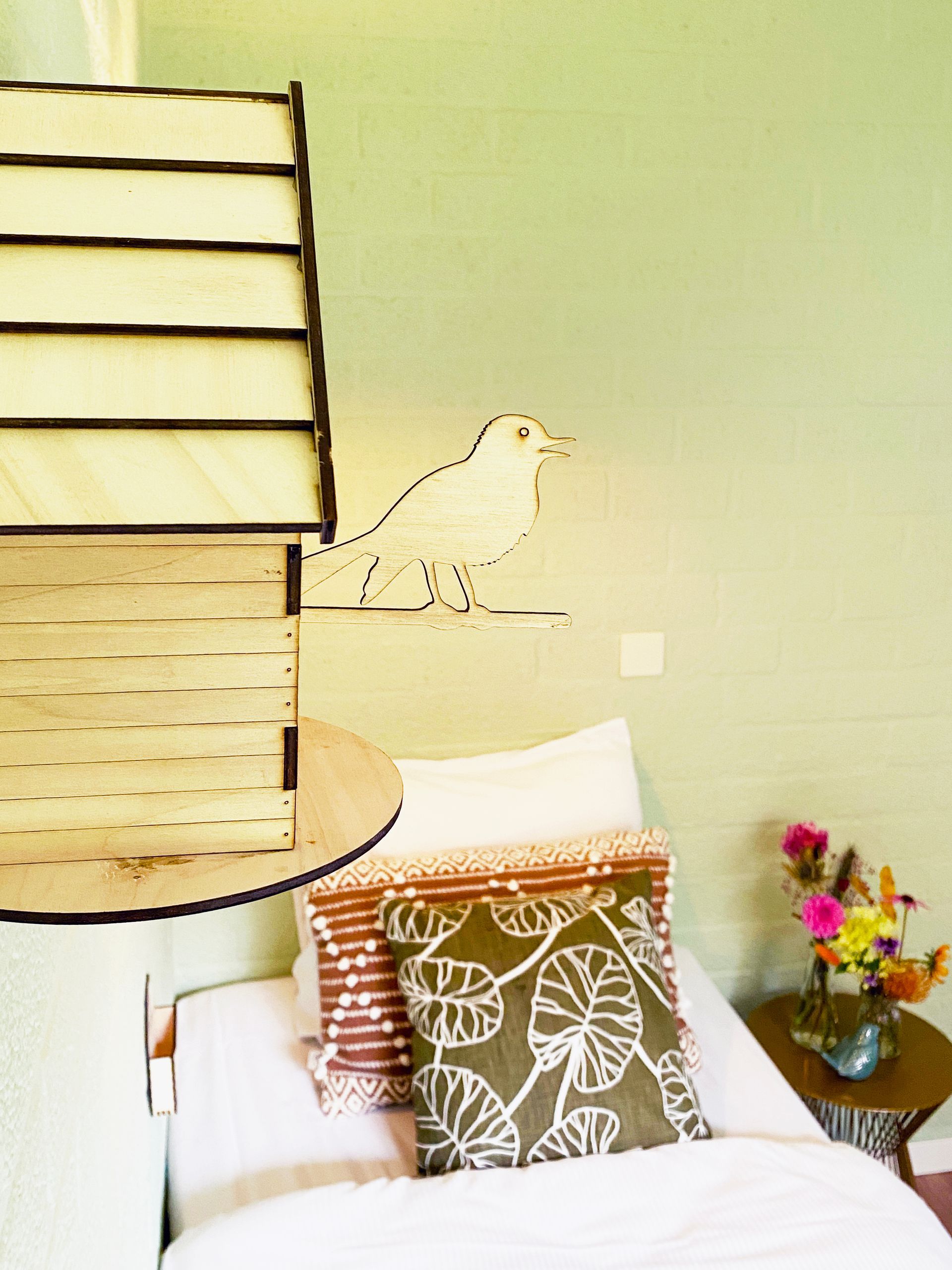 Vogelhuisje en bed met kussens met patronen tegen een lichtgroene muur, klein tafeltje met bloemen.