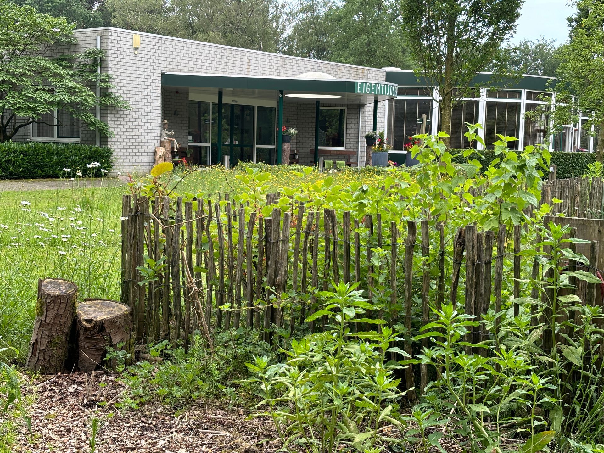 Een laag houten hek omringt een tuin met een gebouw op de achtergrond. Groen vult het tafereel.