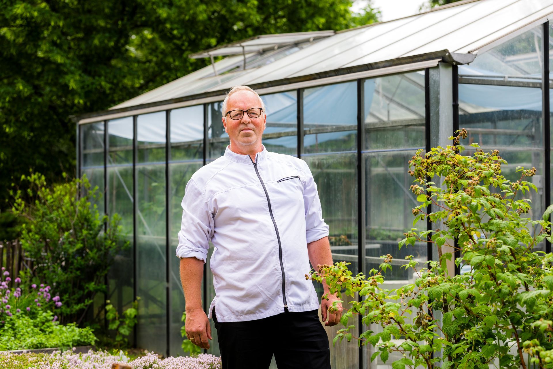 Een man in een wit shirt staat voor een kas; op de voorgrond zijn tuinplanten te zien.