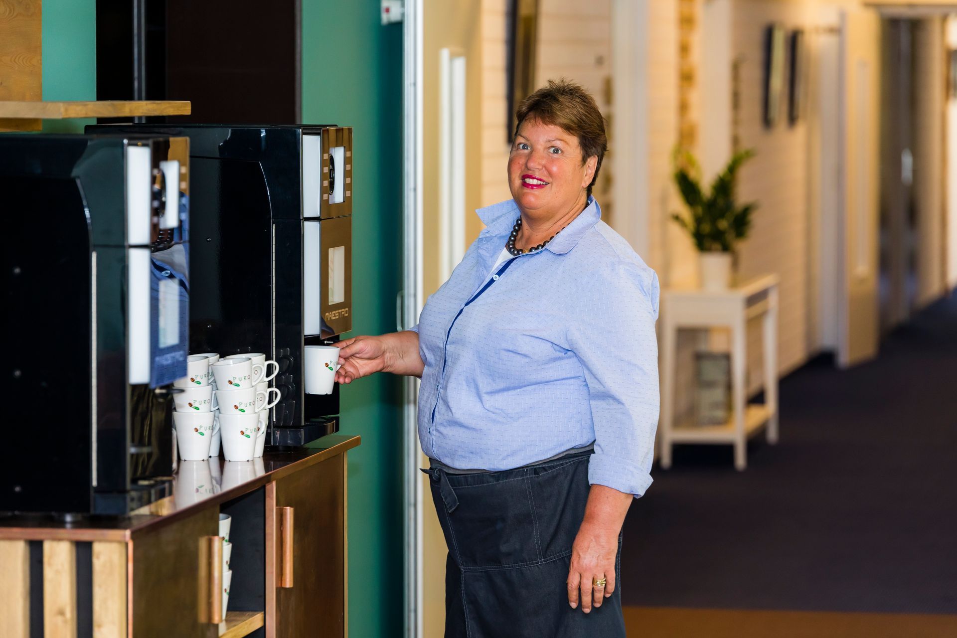 Een vrouw schenkt koffie in bij een koffiezetapparaat in een felverlichte kantoorruimte.