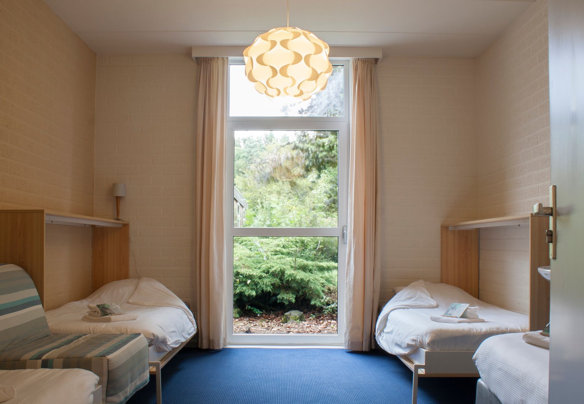 Drie bedden in een kamer met een groot raam dat uitkijkt op bomen, beige muren en een blauw tapijt.