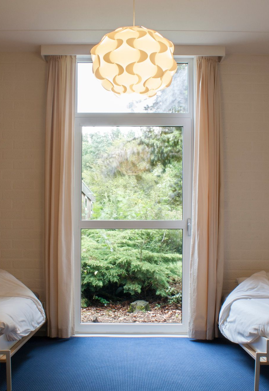 Drie bedden in een kamer met een groot raam dat uitkijkt op bomen, beige muren en een blauw tapijt.