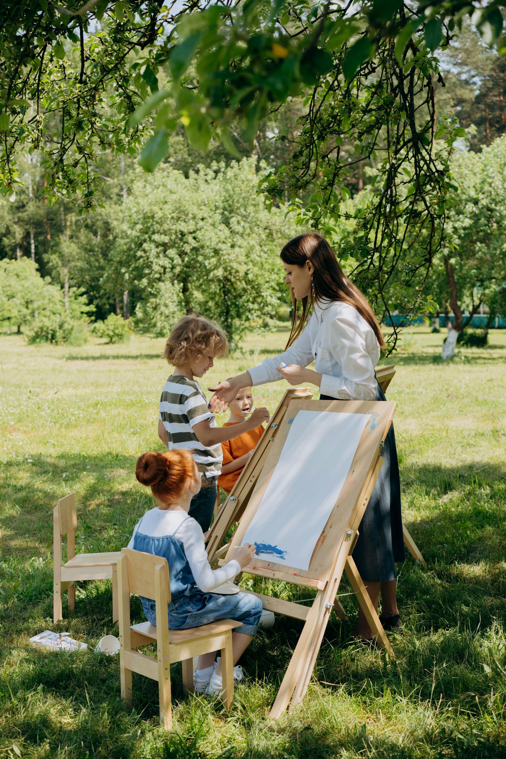 Kinderen en de leerkracht schilderen buiten in een zonnig park aan schildersezels.