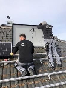 Zwei Arbeiter installieren Solarpaneele auf einem Dach. Einer kniet, der andere steht. Sonnenlicht.