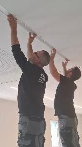 Zwei Personen montieren Deckenleuchten. Beide tragen dunkle Hemden und Arbeitshosen.