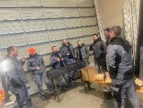 Eine Gruppe Männer grillt im Freien in der Nähe eines Metallgebäudes. Einige tragen graue Arbeitsuniformen, andere Jacken; sie unterhalten sich.