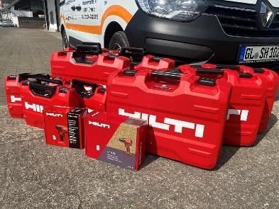 Rote Hilti-Werkzeugkästen und Bohrmaschinenverpackungen vor einem weiß-orangenen Servicefahrzeug auf einer Asphaltfläche.