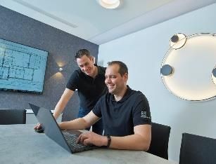 Zwei Männer in schwarzen Hemden arbeiten lächelnd an einem Laptop in einem modernen Büro zusammen.