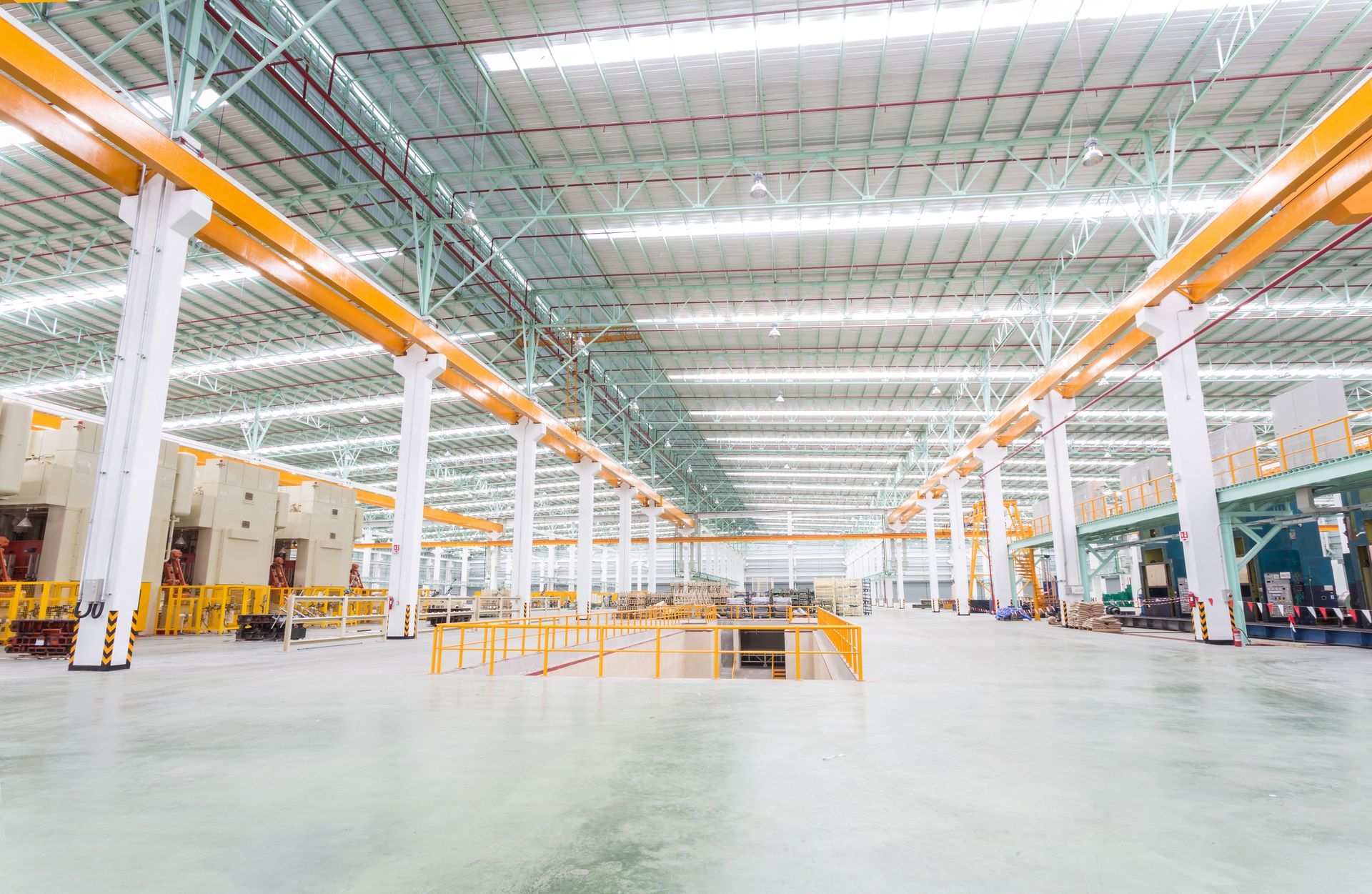 Vue intérieure grand angle d'un vaste hall d'usine industriel lumineux, avec de hauts plafonds et des ponts roulants jaunes.