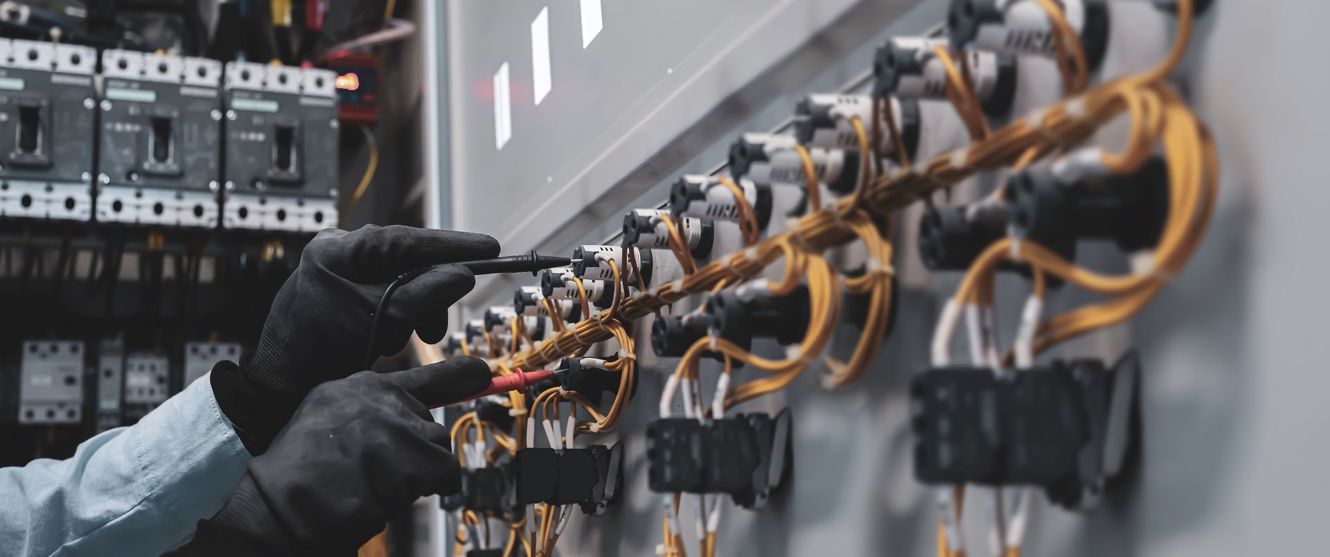 Un électricien portant des gants noirs teste les connexions de câblage sur un tableau électrique complexe à l'aide de multimètres.