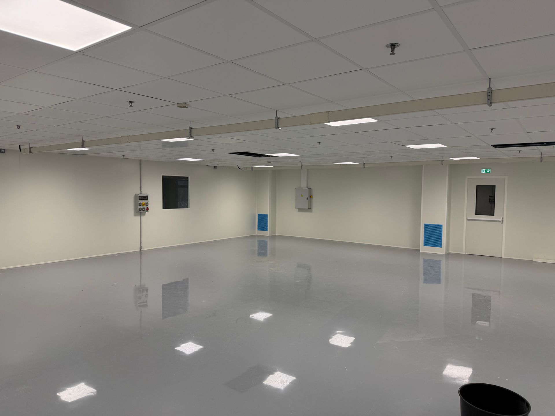 Une grande pièce vide avec un sol gris brillant, des murs blanc cassé, un faux plafond avec des lumières et une porte sur la droite.
