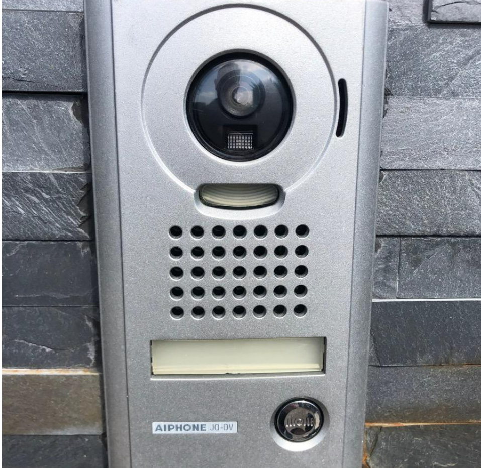 Une sonnette Aiphone métallique avec caméra et haut-parleur intégrés, fixée sur un mur carrelé de pierre gris foncé.